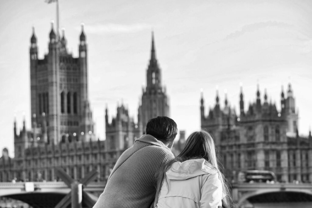 A couple sits together, admiring the iconic Westminster Palace in a black and white photograph.